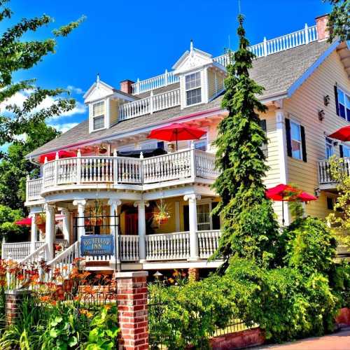 A charming inn with a wraparound porch, surrounded by lush greenery and colorful flowers on a sunny day.