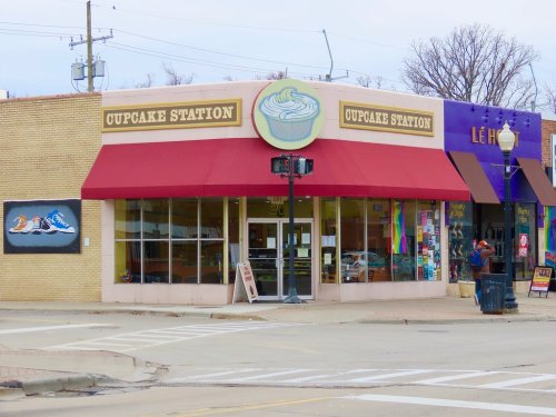 A storefront labeled "Cupcake Station" with a red awning, colorful decorations, and nearby shops on a street corner.