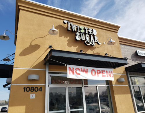 Exterior of Twisted Sugar store with a "NOW OPEN!" banner displayed. Bright yellow wall and clear blue sky.