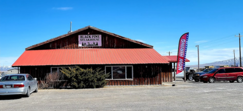 A rustic building with a red roof, labeled "Black Pine Steakhouse," featuring a "Now Hiring" flag outside.