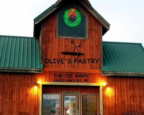 A rustic wooden building with a green roof, featuring a sign for "Olive's Pastry" and "The Pie Barn," decorated for the holidays.