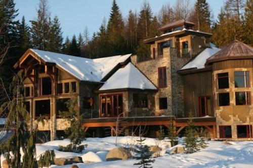 A large, modern cabin with stone and wood features, surrounded by snow and evergreen trees in a winter landscape.