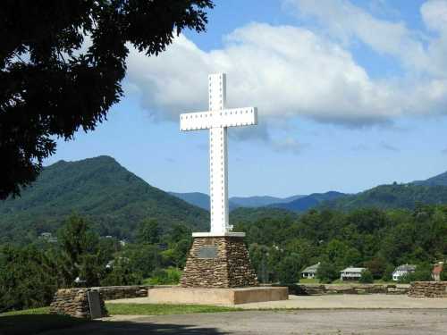 A large white cross stands on a stone base, surrounded by green hills and a blue sky with clouds.