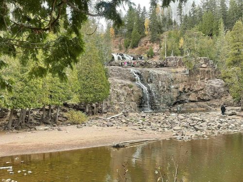 A serene landscape featuring a waterfall cascading over rocks, surrounded by trees and a calm lake in the foreground.