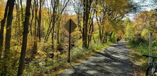 A scenic path lined with trees in autumn colors, with a cyclist in the distance and a creek nearby.