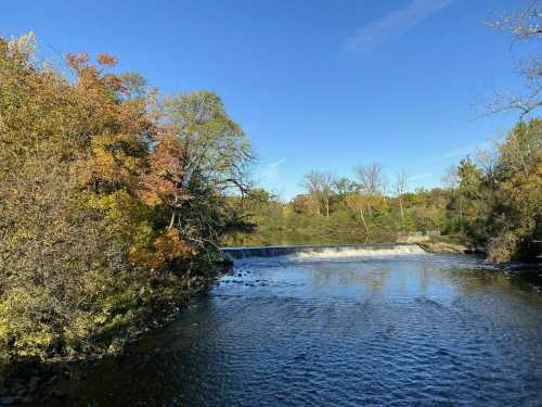 A serene river scene with a small waterfall, surrounded by trees displaying autumn colors under a clear blue sky.