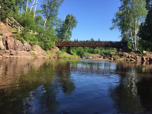 A serene river scene with a wooden bridge, surrounded by lush greenery and clear blue skies.