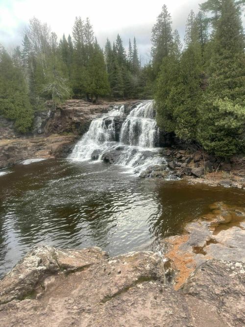 A serene waterfall cascades over rocks into a calm pool, surrounded by lush green trees and a cloudy sky.