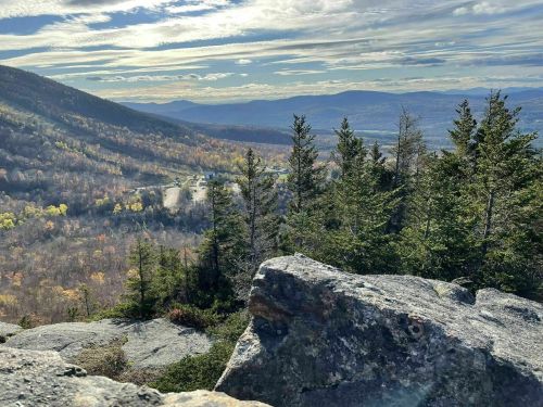 A scenic view of mountains and valleys, with rocky foreground and trees, under a cloudy sky. Autumn colors are visible.