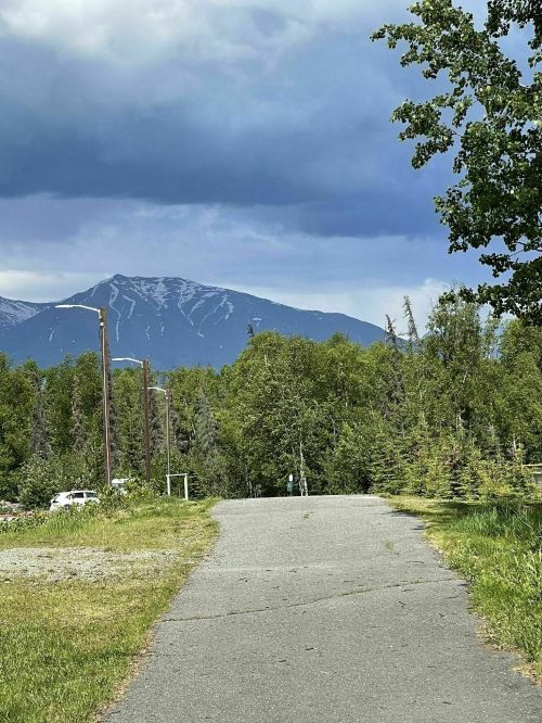 A paved path leads through greenery towards a mountain under a cloudy sky.
