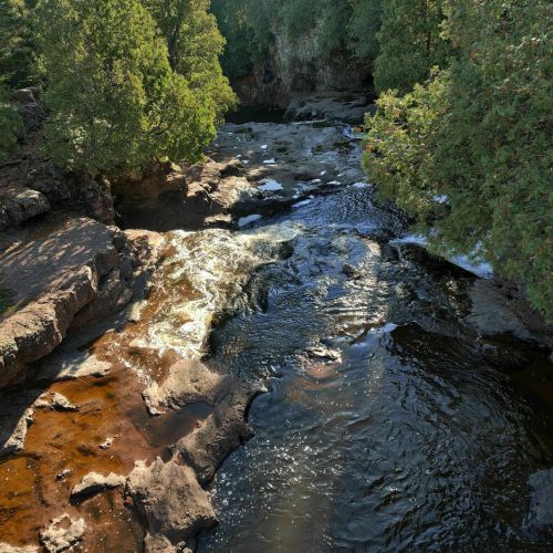 A serene river flows through rocky terrain, surrounded by lush green trees and sunlight filtering through the leaves.