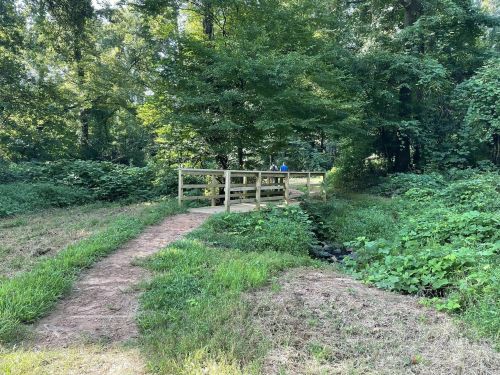 A wooden bridge over a small stream, surrounded by lush greenery and trees in a peaceful outdoor setting.