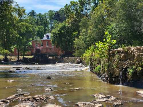 A serene river scene with rocks, flowing water, and a historic red building surrounded by lush greenery.