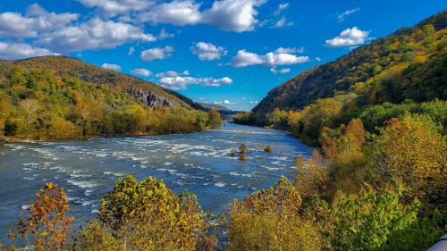 A scenic river flows between colorful autumn hills under a blue sky with fluffy clouds.