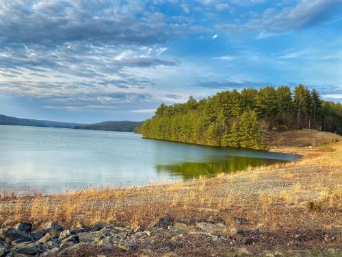 A serene lake surrounded by trees under a partly cloudy sky, with a rocky shoreline and grassy areas.