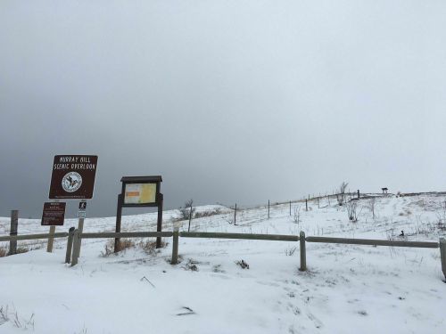 Snow-covered landscape with a sign for Murray Hill Scenic Overlook, under a cloudy sky. Fenced area in the foreground.