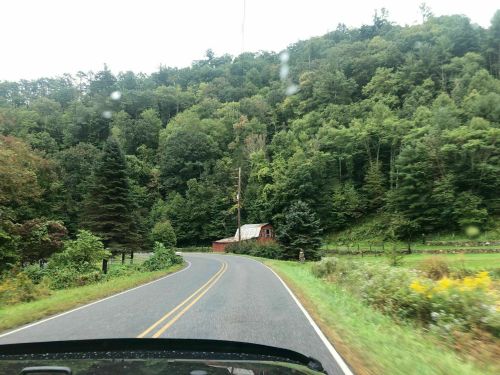 A winding road through lush green trees, with a small red barn visible in the distance. Rain droplets on the windshield.