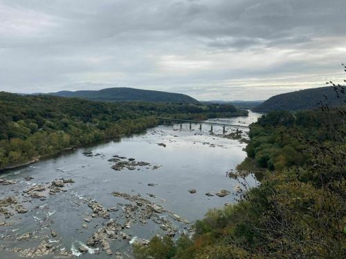 A scenic view of a river winding through green hills, with a bridge crossing in the distance under a cloudy sky.