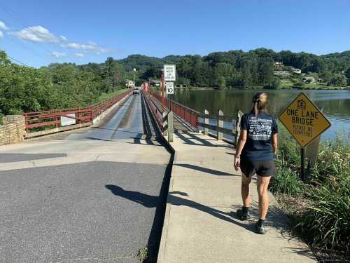 A person walks towards a one-lane bridge over a calm river, surrounded by greenery and blue skies.