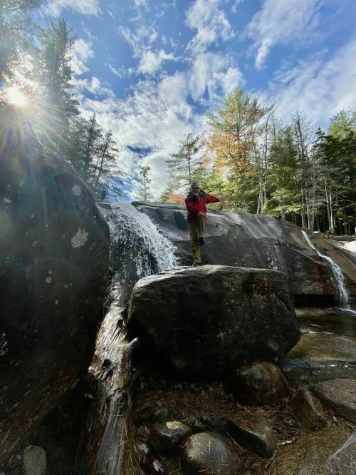 A person in a red jacket balances on a rock near a waterfall, surrounded by trees and a blue sky with clouds.