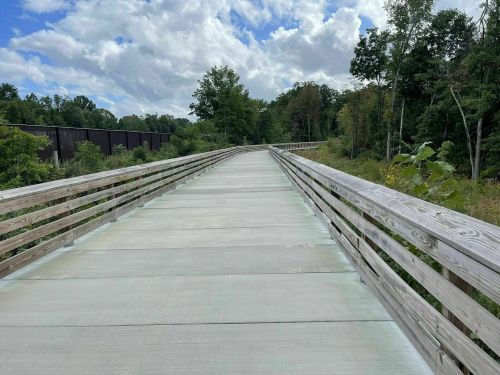 A wooden bridge stretches into the distance, surrounded by greenery and under a partly cloudy sky.