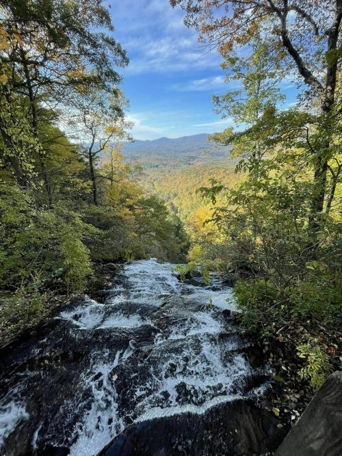 A cascading waterfall flows down rocky terrain, surrounded by vibrant autumn foliage and distant mountains under a blue sky.