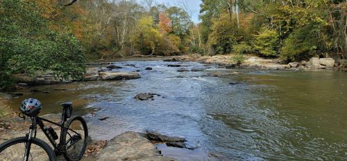 A bike rests on a rocky riverbank, surrounded by trees with autumn foliage and a gently flowing river.