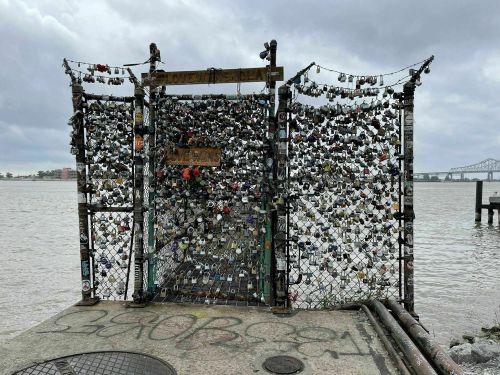 A fence covered in padlocks stands by a river, with a cloudy sky in the background.