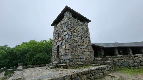 A stone tower stands amidst greenery under a cloudy sky, with steps leading up to it.