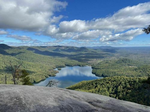 A panoramic view of a lush green valley with a serene lake, surrounded by mountains under a partly cloudy sky.