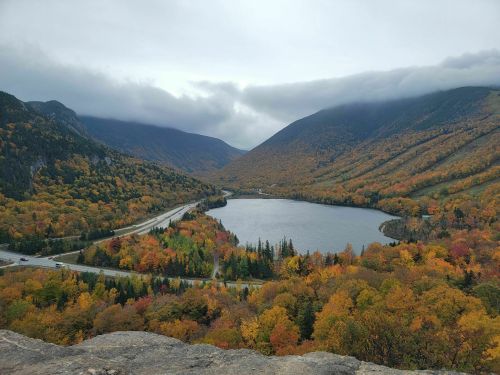 A scenic view of a lake surrounded by autumn foliage and mountains under a cloudy sky.