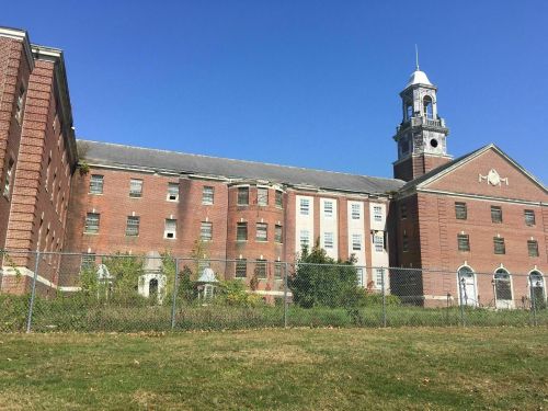 Abandoned brick building with a clock tower, surrounded by a fence and overgrown grass under a clear blue sky.