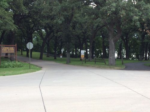 A park entrance with a sign for Lower Oak Park Access, surrounded by trees and a winding road leading to a lake.