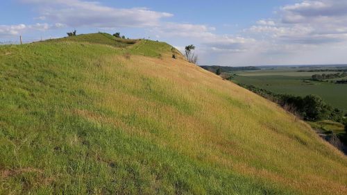 A grassy hillside with gentle slopes, under a blue sky with scattered clouds, overlooking a green landscape.