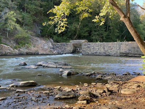 A serene river scene with a stone dam, surrounded by trees and rocky banks under a clear sky.