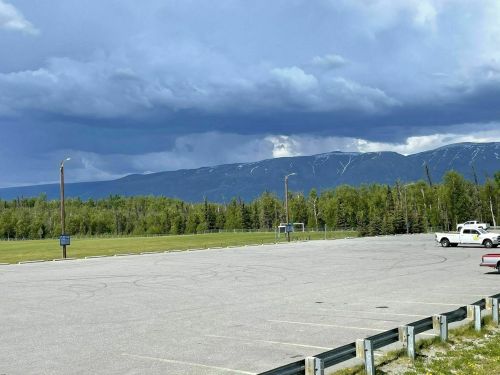 A wide view of an empty parking lot with mountains and dark clouds in the background, surrounded by trees.