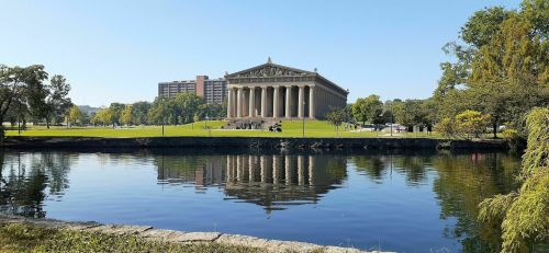 A large classical building with columns reflects in a calm lake, surrounded by green trees and a clear blue sky.