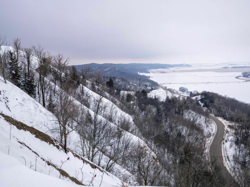 Snow-covered hills and a winding road stretch through a winter landscape under a cloudy sky.