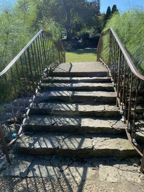 Stone steps lead up to a pathway, flanked by greenery and a railing, under a clear blue sky.