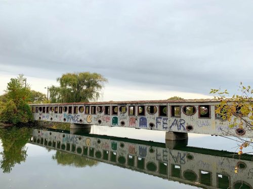 A graffiti-covered bridge spans a calm river, reflecting the cloudy sky and surrounding trees.