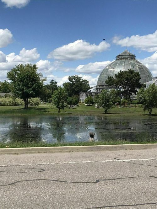 A large glass conservatory surrounded by greenery and a reflective pond under a blue sky with fluffy clouds.