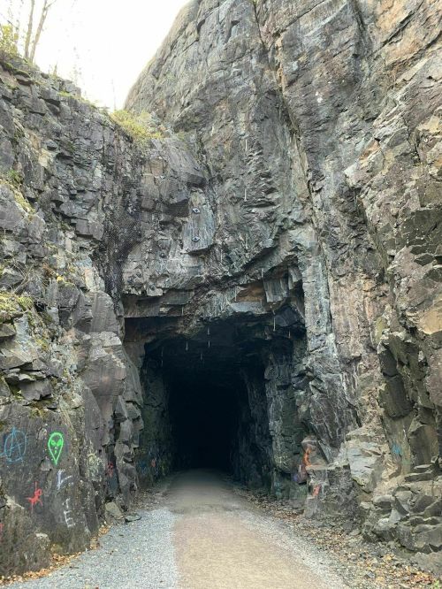 A dark cave entrance surrounded by rocky cliffs, with colorful graffiti on the stone walls and a gravel path leading inside.