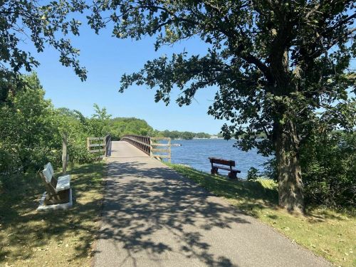 A peaceful pathway leads to a bridge over a lake, surrounded by trees and benches on a sunny day.