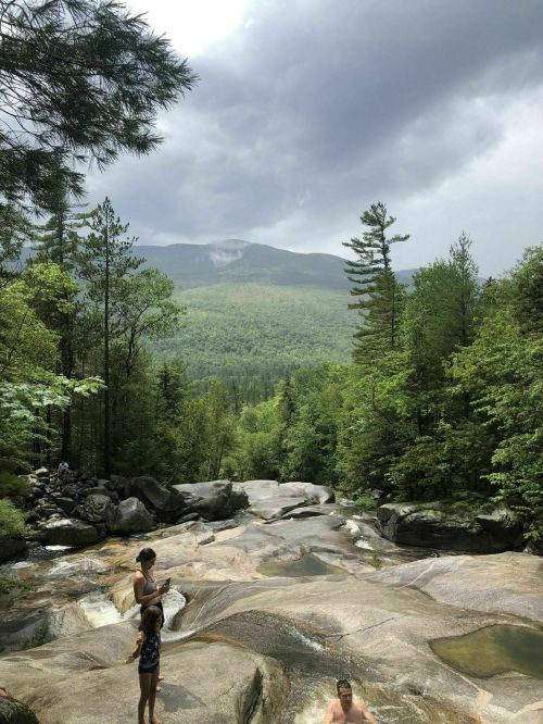 A scenic view of a rocky river surrounded by lush green trees and mountains under a cloudy sky. Two people are near the water.
