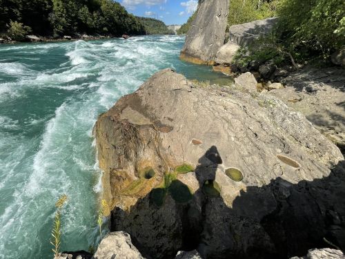 A rocky riverbank with rushing turquoise water and a shadow cast on the stone, surrounded by greenery and cliffs.