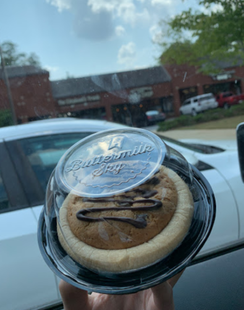 A hand holding a cookie in a clear container, with chocolate drizzle, against a car window view.