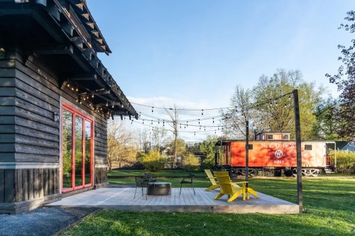 A modern cabin with a deck and yellow chairs, surrounded by greenery and a vintage train car in the background.