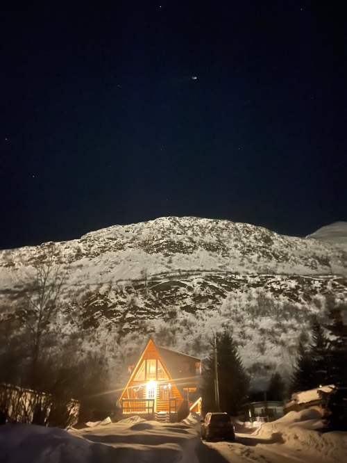A cozy cabin illuminated at night, surrounded by snow and mountains under a starry sky.