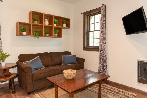 Cozy living room with a brown sofa, wooden coffee table, wall shelves with plants, and a TV mounted on the wall.