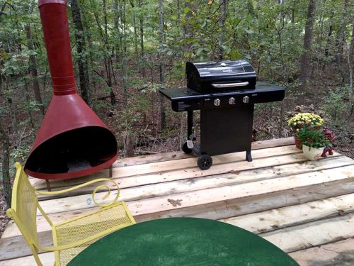 A backyard patio with a black grill, red outdoor fireplace, yellow chair, and potted flowers surrounded by trees.
